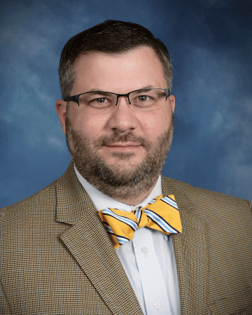 Professional portrait of a man with a beard wearing glasses and a bow tie, dressed in a patterned suit jacket, against a blue background.